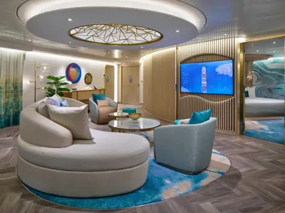 Stilvoller Wohnbereich in einer Suite der Utopia of the Seas, mit runder Couch, Sesseln, TV und modernem Design. Blick ins angrenzende Schlafzimmer.