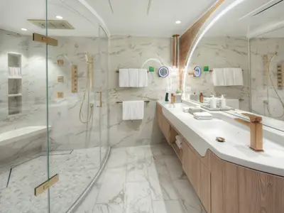 Helles, luxuriöses Bad mit begehbarer Dusche und Sitzbank. Doppelwaschtisch mit goldenen Armaturen und großem, beleuchtetem Spiegel. Marmorfliesen und helle Holzmöbel dominieren das elegante Design.