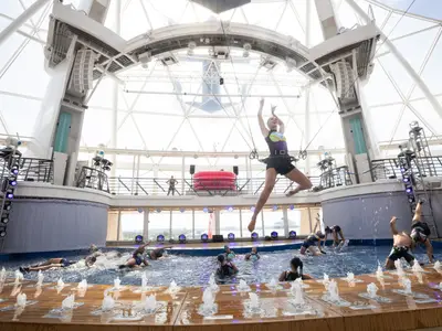 Akrobatik-Wassershow auf der Utopia of the Seas. Eine Artistin schwebt hoch über einem Pool mit Akrobaten und Wasserfontänen. Die Szene spielt unter einem Glasdach.