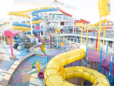 Das bunte Aqua-Park-Deck der Utopia of the Seas zeigt Wasserrutschen in Gelb, Blau und Orange, sowie einen interaktiven Splash-Bereich mit verspielten Elementen, teilweise noch im Bau.