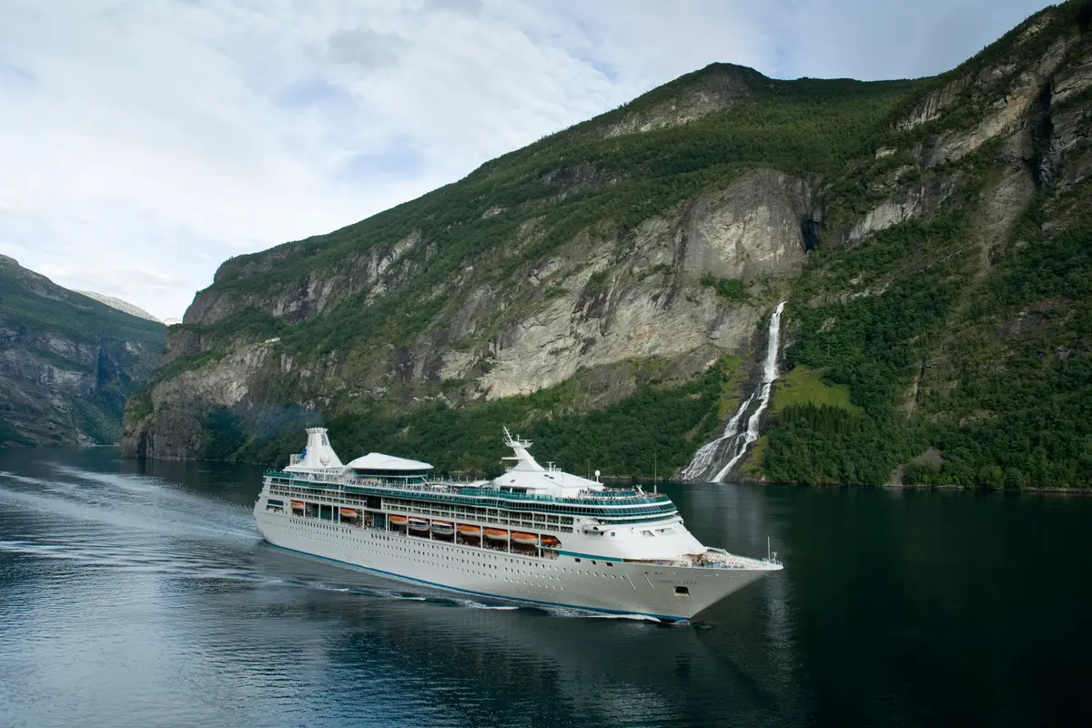Die Vision of the Seas, ein weißes Schiff, fährt durch einen Fjord. Steile, grüne Berge umgeben das Schiff, rechts fällt ein Wasserfall hinab.