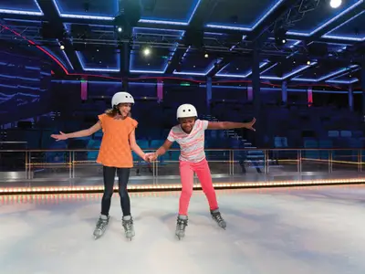 Zwei Kinder, lachend und mit Helmen, Schlittschuhen, halten Händchen beim Eislaufen auf der Voyager of the Seas. Ein Erwachsener ist im Hintergrund der blauen Eisarena zu sehen.