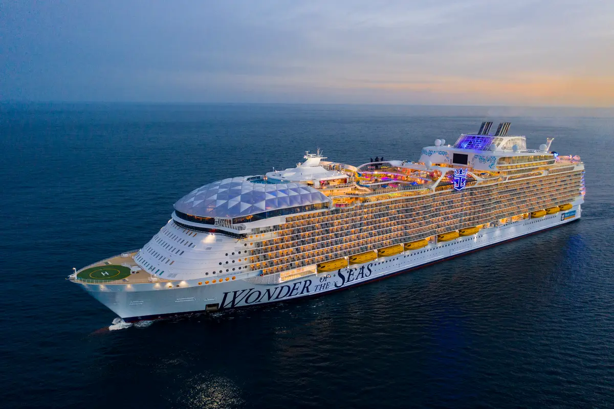 Die große Wonder of the Seas auf dem Meer bei Abenddämmerung. Die erleuchteten Balkonkabinen und bunten Oberdecks sind gut sichtbar. Am Bug befindet sich ein Helipad.