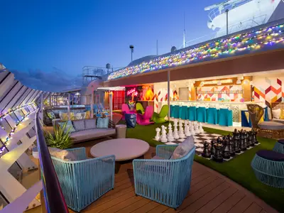 Abendliche Aufnahme eines bunten Unterhaltungsdecks der Wonder of the Seas. Zu sehen sind eine Bar, verschiedene Sitzbereiche, ein großes Schachspiel auf Kunstrasen und festliche Lichterketten.