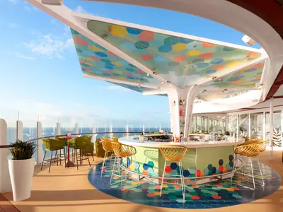Ein lebendiges Außen-Barbistro auf der Wonder of the Seas unter einem farbenfrohen Dach. Gelbe Barhocker umgeben die Theke, im Hintergrund glänzt das Meer.