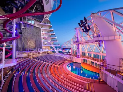 Das beleuchtete AquaTheater-Deck der Wonder of the Seas am Abend. Zu sehen sind Sitzreihen, der Pool, eine Kletterwand und pinke Wasserrutschen.