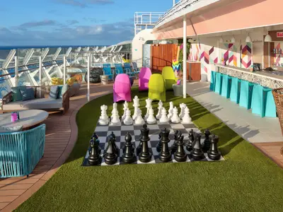 Ein weitläufiger Außendeckbereich der Wonder of the Seas mit einem großen Schachspiel auf Kunstrasen, farbigen Stühlen, einer Bar mit türkisen Hockern und bequemen Lounge-Möbeln mit Meerblick.