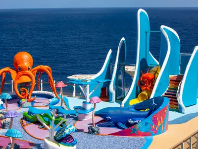 Ein lebhafter Kinder-Wasserspielplatz auf dem Oberdeck der Wonder of the Seas. Zu sehen sind eine große orange Krake, ein blauer Wal, Rutschen, sowie bunte Pilz- und Unterwasserfiguren mit dem weiten Ozean im Hintergrund.
