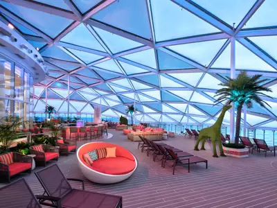 Das überdachte Solarium-Deck der Wonder of the Seas zeigt eine Bar, Lounge-Sessel, Liegen, Palmen und eine Giraffen-Skulptur. Meerblick im Hintergrund.