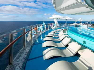Blick auf das Oberdeck der Wonder of the Seas: Ein Poolbereich mit Liegestühlen im flachen Wasser und Glasgeländer zum offenen Meer unter blauem Himmel.
