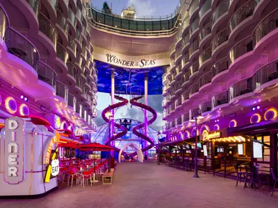 Blick auf die hell erleuchtete Boardwalk-Promenade der Wonder of the Seas bei Dämmerung, gesäumt von Restaurants, Bars und Balkonkabinen. Im Hintergrund sind hohe Wasserrutschen zu sehen.