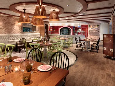 Ein helles Restaurant an Bord der Wonder of the Seas. Es zeigt Holztische mit hellgrünen und dunklen Stühlen, eine Ziegelwand, einen Pizzaofen und eine Theke.