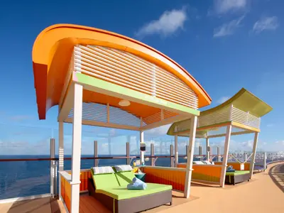 Bunte Cabanas des '10. Palms Park' auf dem sonnigen Oberdeck der Wonder of the Seas. Grüne Liegen mit blauen Handtüchern und weitem Meerblick unter blauem Himmel sind sichtbar.