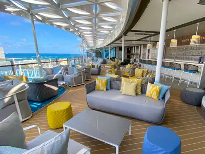 Das helle Außendeck der Wonder of the Seas zeigt eine Lounge mit grauen Sofas, gelben und blauen Kissen und einer Bar. Im Hintergrund sind das Meer und die Küste sichtbar, überdacht von einem modernen Sonnenschutz.