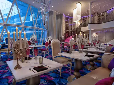 Innenaufnahme eines luxuriösen Restaurants der Wonder of the Seas mit Tischen, Kerzen, buntem Teppich, einer prächtigen Treppe und großen Fenstern mit Blick nach draußen.