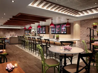 Innenansicht eines stilvollen Restaurants/Barbereichs auf der Wonder of the Seas mit Holztischen, Stühlen, einer langen Bar, beleuchteten Flaschenregalen, roten Hängelampen und Ziegelwänden.