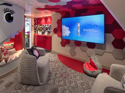 Themensuite der Wonder of the Seas mit Hai-Bett und roter Küchenzeile. Rote und graue Sechseckpaneele an Wänden und Decke. Sitzgelegenheiten.