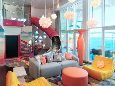Blick in einen bunten Familienbereich der Wonder of the Seas. Eine rote Röhrenrutsche, eine Wendeltreppe, Sitzgelegenheiten und Meerblick durch große Fenster sind dominant.