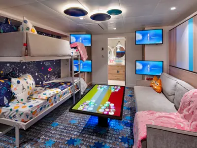 Kinderkabine der Wonder of the Seas. Links Stockbett, mittig interaktiver Tisch, rechts Couch, mehrere Bildschirme. Design mit Weltraum-Motiven.