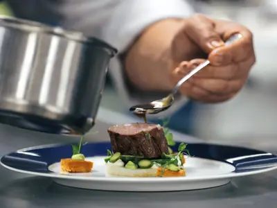 Nahaufnahme eines Koches, der Sauce mit einem Löffel kunstvoll über ein arrangiertes Gourmet-Fleischgericht träufelt.
