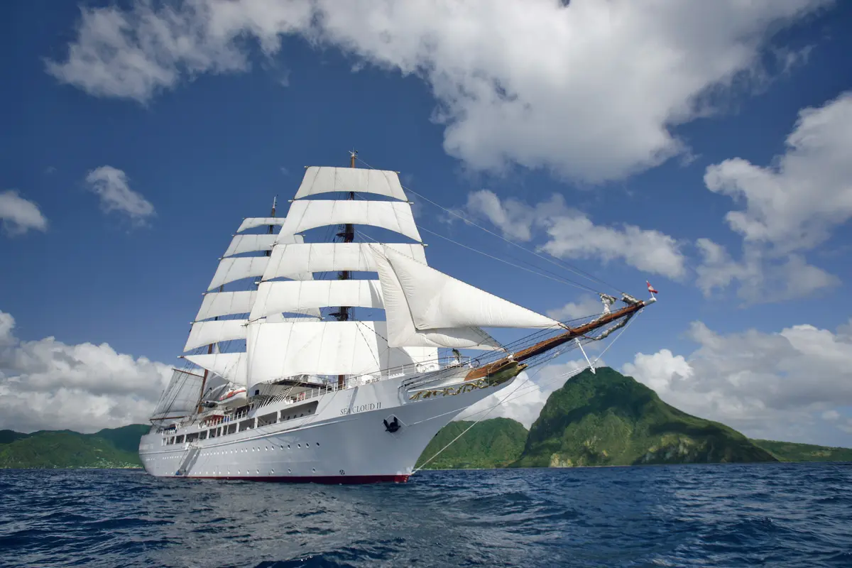 Der Großsegler Sea Cloud II gleitet mit gesetzten weißen Segeln auf blauem Meer an grünen Bergen vorbei.