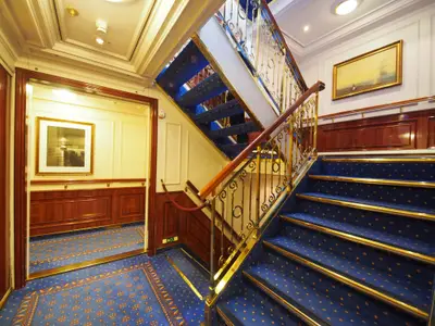 Klassisches Treppenhaus an Bord der Sea Cloud Ii mit blau-goldenen Teppichen, Holzverkleidung und goldener Messingbrüstung.