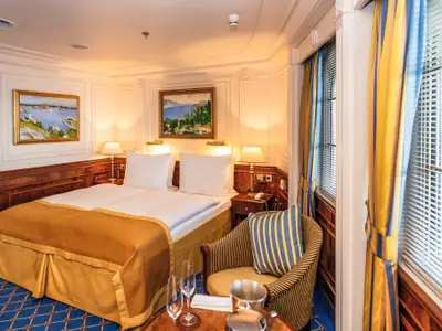 Elegante Außenkabine an Bord der Sea Cloud II mit großem Doppelbett, Nachttischlampen, Gemälden und einem gemütlichen Sessel.