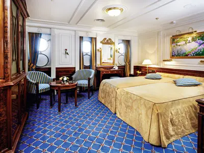 Elegante Deluxe-Außenkabine an Bord der Sea Cloud Ii mit Doppelbett, blauen Sesseln und maritimen Bullaugen.