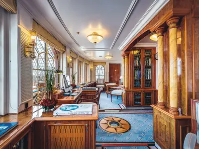 Elegante Bibliothek an Bord der Sea Cloud II mit Holzregalen, Schreibtischen, Lampen und blauem Teppich mit Windrose.