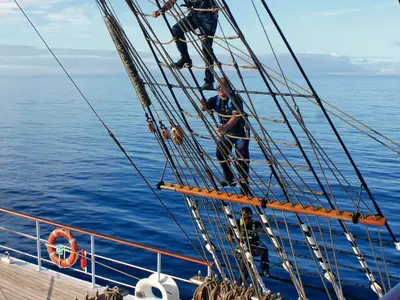 Zwei Personen klettern gesichert in den Takelagen und Seilen an Bord der Sea Cloud Ii mit Blick auf den Ozean.