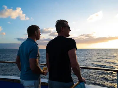 Zwei Männer stehen am Abend an der Holzreling an Bord der Sea Cloud Ii und blicken auf den Sonnenuntergang über dem Meer.