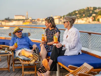 Drei Gäste sitzen entspannt mit Cocktails auf Sonnenliegen an Deck der Sea Cloud II und genießen den Blick auf die Küste.