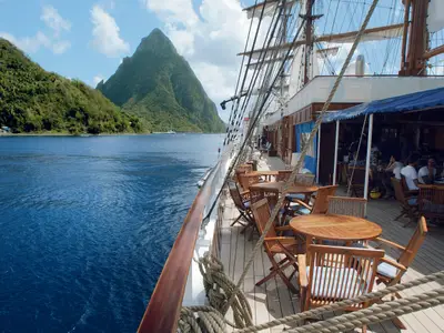 Blick über das Holzdeck der Sea Cloud II mit Tischen und Stühlen auf das blaue Meer und steile grüne Berge.