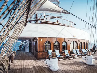 Blick über das sonnige Holzdeck der Sea Cloud II mit blauen Liegestühlen, Masten und Seilen vor blauer Seekulisse.
