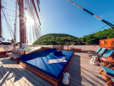 Blick auf das Holzdeck der Sea Cloud II mit blauen Liegekissen, Masten und grünen Hügeln im Hintergrund.