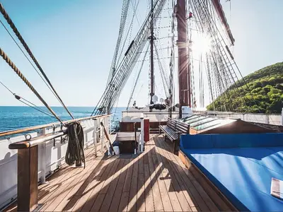 Blick auf das Holzdeck der Sea Cloud Ii mit blauen Liegekissen, Segelmasten und Aussicht auf das Meer und grüne Küste.