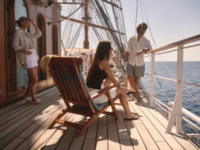 Drei Gäste entspannen auf dem Holzdeck des Segelschiffs Sea Cloud Spirit mit Blick auf das offene Meer bei strahlendem Sonnenschein.