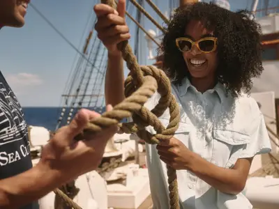 Eine lächelnde Frau mit Sonnenbrille knüpft gemeinsam mit einem Mann einen dicken Knoten aus Tauwerk an Bord der Sea Cloud Spirit.