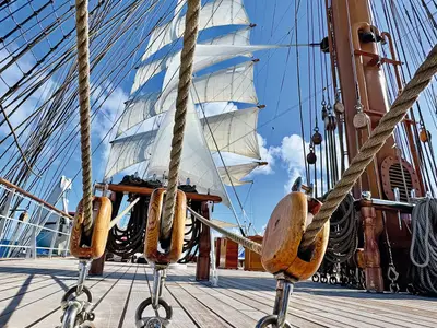 Blick über das Holzdeck der Sea Cloud Spirit hinauf zu den gespannten weißen Segeln, Masten und Seilen bei blauem Himmel.