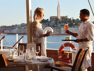 Ein Kellner reicht einer eleganten Frau einen Drink auf dem Deck der Sea Cloud Spirit vor einer Küstenstadt.