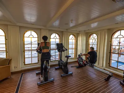 Zwei Gäste trainieren an Fitnessgeräten im hellen Fitnessraum an Bord der Sea Cloud Spirit mit großen Bogenfenstern aufs Meer.