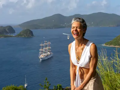 Eine fröhlich lachende Frau genießt die Aussicht auf eine karibische Bucht mit dem Segelschiff Sea Cloud Spirit im Meer.