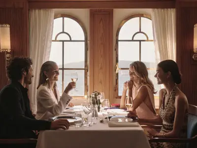 Vier elegant gekleidete Gäste stoßen im Restaurant an Bord der Sea Cloud Spirit mit Weißwein an einem gedeckten Tisch an.