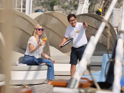 Eine Frau mit Sonnenbrille genießt auf dem Sonnendeck der Sea Cloud Spirit einen Cocktail, während ein Crewmitglied lächelt.