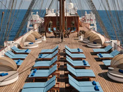 Blick über das hölzerne Sonnendeck der Sea Cloud Spirit mit blauen Liegestühlen, Daybeds und Masten vor dem Meer.