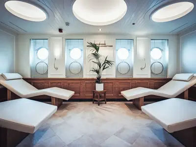 Ein heller Wellness-Ruheraum mit vier weißen Liegen, Holzvertäfelung und Bullaugen an Bord der Sea Cloud Spirit.