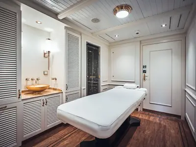 Heller Massageraum an Bord der Sea Cloud Spirit mit weißer Liege, goldener Waschschüssel und Dusche im Hintergrund.