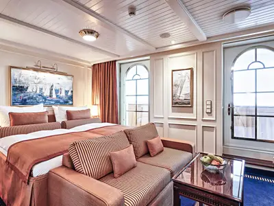 Blick in eine edle Junior Veranda Suite an Bord der Sea Cloud Spirit mit gestreiftem Sofa, großem Bett und Segelschiff-Kunst.