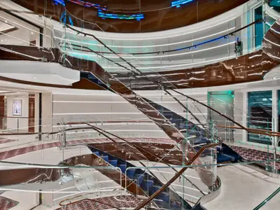 Blick auf die kunstvoll geschwungene Treppe mit glänzenden Holzelementen und Glasgeländern im Atrium an Bord der Seabourn Encore.