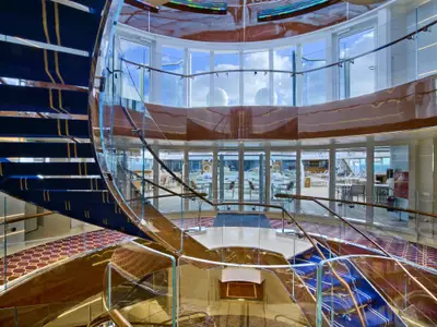 Blick auf die geschwungene Treppe mit blauem Teppich und Glasgeländer im hellen Atrium an Bord der Seabourn Encore.